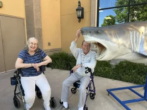 An elderly woman and man with walkers pose next to a large model of a shark at the Norcal Aquarium; the man is playfully placing his hand inside the shark’s mouth during their Wildlife Adventures outing.