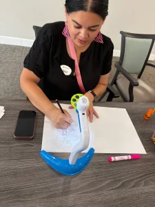 A woman sits at a table drawing Nurse Appreciation Cards on white paper with a pencil, using a blue and white drawing tool. A phone and a marker are on the table.