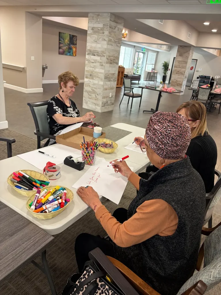 Three women sit at a table in a communal room, creating colorful Nurse Appreciation Cards with markers, paper, and baskets of supplies.