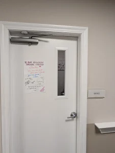 A closed white door with a small window and a handwritten note taped to it, next to a wall sign labeled "Office," features colorful Nurse Appreciation Cards displayed inside.