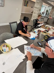 Several people sit at a table creating Nurse's Day Cards and thank-you notes with markers and art supplies in a communal indoor setting.