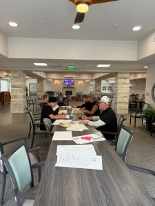 Five adults sit at a long table in a spacious, well-lit room, creating colorful Nurse's Day Cards with papers and markers spread out in front of them.