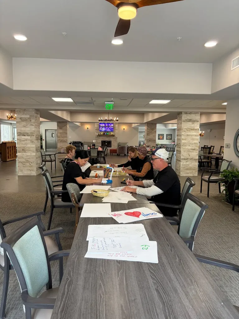 Five adults sit at a long table in a spacious, well-lit room, creating colorful Nurse's Day Cards with papers and markers spread out in front of them.