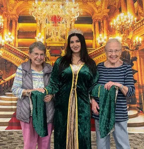 Three women, one dressed as Queen Esther in a green medieval gown and crown, stand before a grand staircase backdrop.