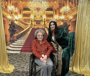 An elderly woman in a wheelchair sits beside a younger woman dressed as Queen Esther in a green gown and crown, with a grand staircase and chandeliers behind them, evoking a sense of jubilee and regal celebration.