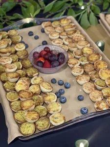 A tray with mini quiches arranged to form the letters "M" and "D," a bowl of mixed berries in the center, and scattered blueberries on the tray—perfect for a Jubilee celebration fit for Queen Esther.