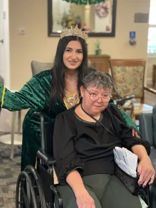 A woman in a green dress and crown, evoking Queen Esther at a Jubilee, stands smiling behind an older woman sitting in a wheelchair, holding papers in her lap, in a room with chairs and framed pictures.