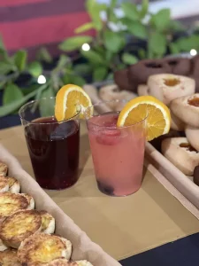 Two drinks, one red and one pink with fruit garnishes, are placed on a table next to trays of assorted pastries, set against greenery—a festive spread inspired by Queen Esther’s Jubilee.