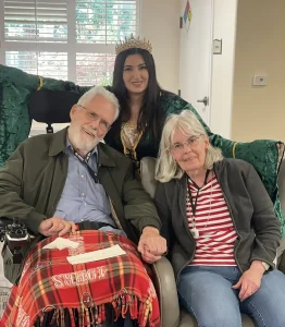 An elderly man in a wheelchair and an elderly woman sit holding hands, with a young woman dressed as Queen Esther, wearing a crown and green robe, standing behind them in what appears to be a Jubilee celebration.