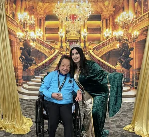 A woman in a green and gold Queen Esther costume poses beside an older person in a wheelchair, with a grand staircase and chandelier creating a regal Jubilee backdrop behind them.