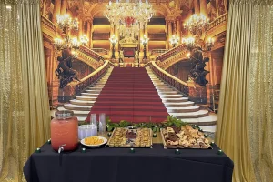 A table with snacks, pastries, cups, and a drink dispenser is set in front of an ornate staircase with chandeliers and golden lighting, evoking the elegance of Queen Esther’s Jubilee celebration.