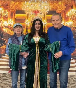 Three people stand in front of a palace-themed backdrop; the woman in the center, dressed as Queen Esther in a green medieval-style dress, is flanked by an older woman and man in casual clothes, celebrating a festive jubilee.