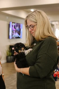 A woman with blonde hair and glasses holds a small black and brown Doodle puppy indoors, looking at it with a thoughtful expression—perhaps considering the joys of raising Doodle puppies from Ranch House Doodles.