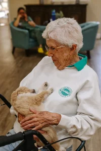 An elderly woman with short gray hair and glasses sits indoors, holding a small, light-colored Doodle puppy in her arms. A person is seated in the background.