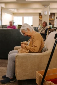 An elderly woman sits on a beige armchair, smiling and holding a doodle puppy in her lap, with two other seniors visible in the background of a well-lit room.