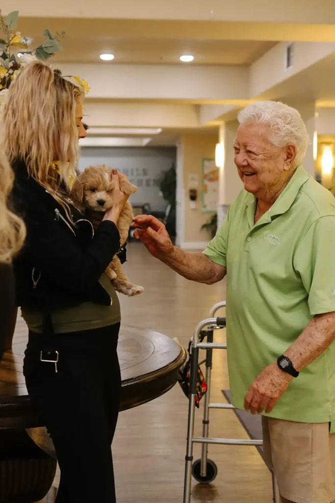 An elderly person in a green shirt smiles and reaches to pet a small Doodle puppy being held by a woman indoors. A walker is nearby, adding warmth to this sweet moment inspired by Ranch House Doodles.
