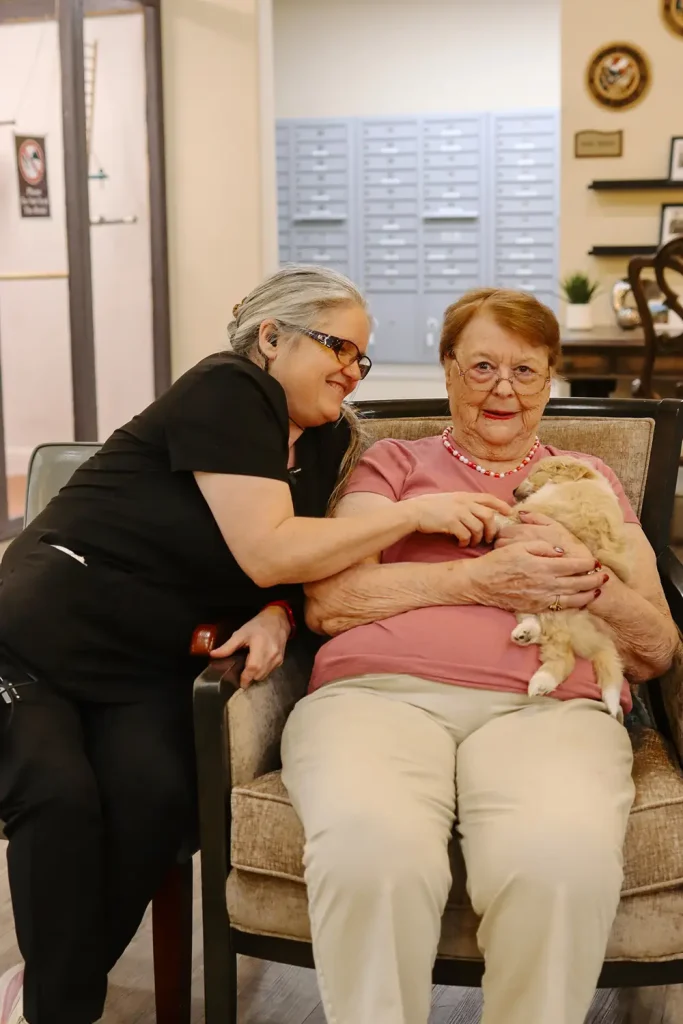 An elderly woman sits on a chair holding a small dog from Ranch House Doodles, while another woman in scrubs sits beside her, smiling and petting the dog indoors.