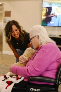 An elderly woman in a wheelchair holds a small Ranch House Doodles pup while a younger woman stands nearby, smiling. A television is on in the background.