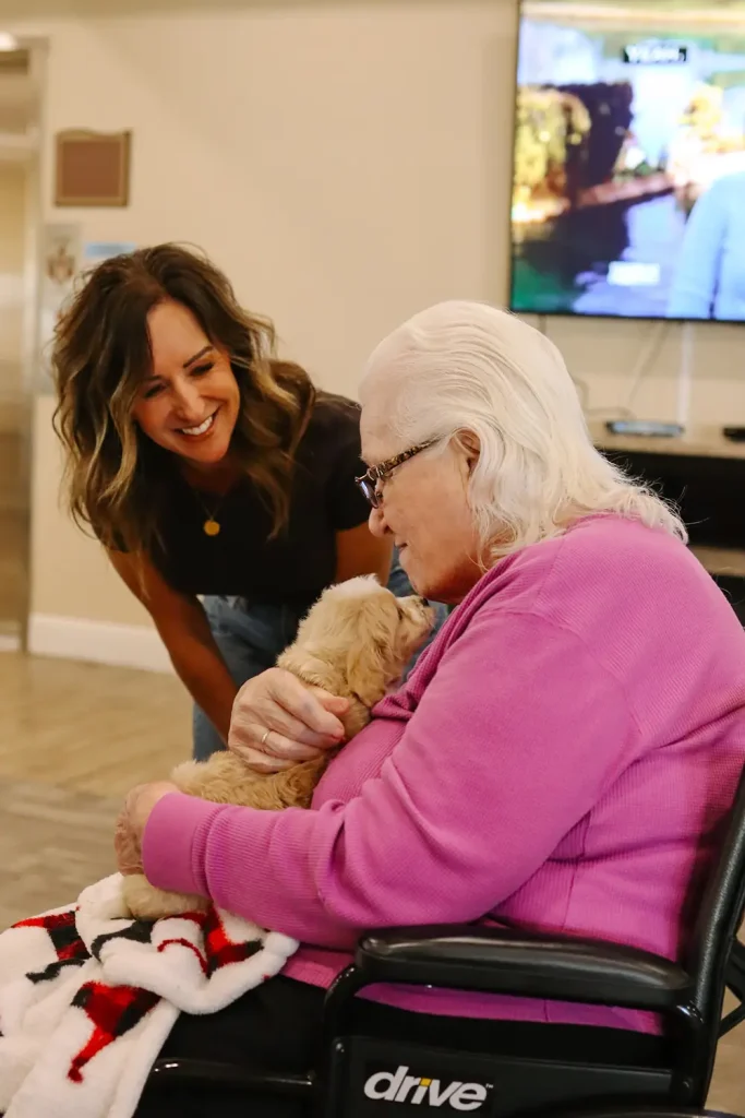 An elderly woman in a wheelchair holds a small Ranch House Doodles pup while a younger woman stands nearby, smiling. A television is on in the background.