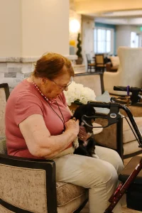 An elderly woman sits in an armchair, petting a small black and white Ranch House Doodles dog in her lap, with a walker beside her in a well-lit room.