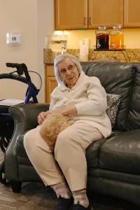 Elderly woman sitting on a couch with a small Doodle puppy on her lap, walker nearby, and kitchen area with beverage dispensers in the background—courtesy of Ranch House Doodles.