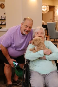An elderly woman in a wheelchair holds a small dog from Ranch House Doodles while a man stands beside her, both looking at the camera indoors.