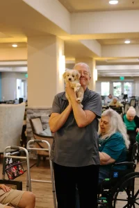 An older adult stands indoors, smiling and holding a small doodle puppy close to their face; other seniors are seated in the background.
