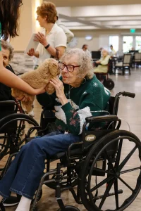 An elderly woman in a wheelchair pets a small dog, possibly one of the adorable Doodle puppies from Ranch House Doodles, held by another person in a care facility, with others present in the background.
