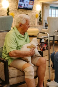 An elderly man with a bandaged knee sits in a chair, holding a small white Ranch House Doodles dog on his lap. A walker with a floral bag rests beside him in the bright room.