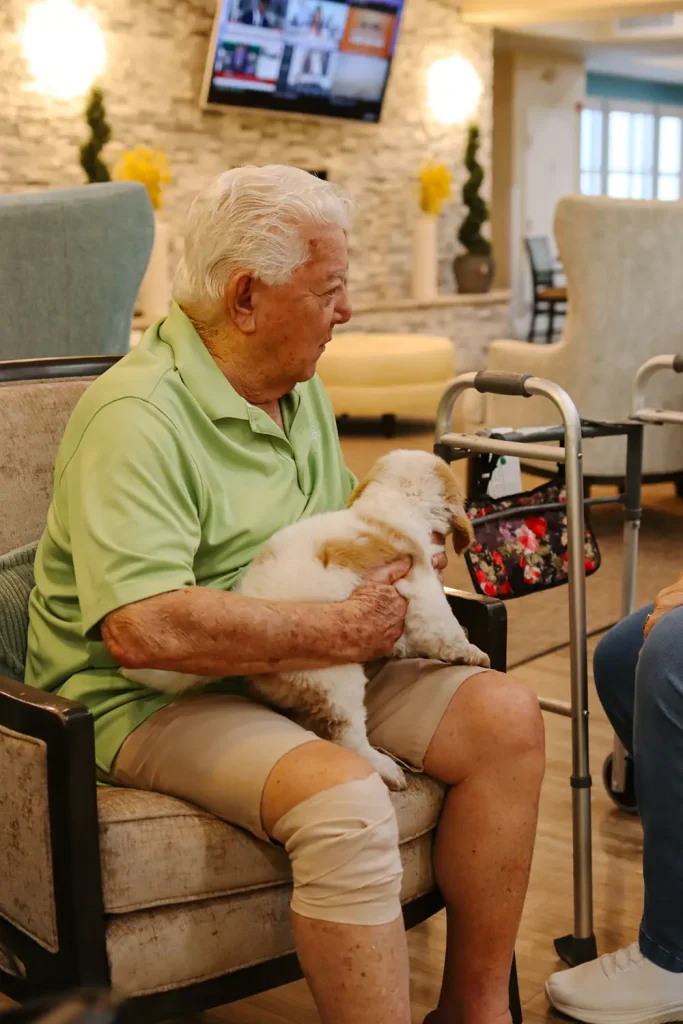 An elderly man with a bandaged knee sits in a chair, holding a small white Ranch House Doodles dog on his lap. A walker with a floral bag rests beside him in the bright room.