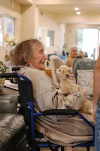 An elderly woman in a wheelchair smiles while holding a small Ranch House Doodles dog on her lap in a bright, communal room with other people in the background.