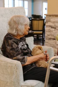 An elderly woman with gray hair sits in an armchair, holding a small Ranch House Doodles dog on her lap. A walker is positioned beside her.