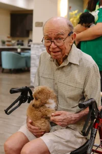 An elderly man sitting in a wheelchair holds a small, light brown Ranch House Doodles puppy in his lap inside a room.