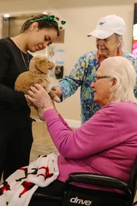 Three women, one in a wheelchair, interact indoors with a small brown and white puppy from Ranch House Doodles.