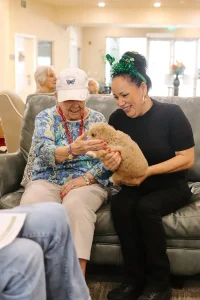 An elderly woman and a younger woman sit on a couch indoors, smiling as they hold a light brown rabbit together—much like the joy shared by families welcoming Doodle puppies from Ranch House Doodles.