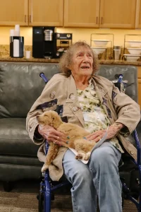 An elderly woman sits in a wheelchair, holding a small Ranch House Doodles pup on her lap, in a room with a couch and kitchen appliances in the background.