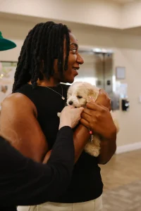 A woman holds a small, light-colored puppy while another person extends a hand to pet the adorable Doodle puppy indoors—just one of many loving companions from Ranch House Doodles.