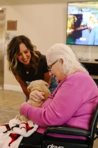 An elderly woman in a wheelchair holds a small Ranch House Doodles puppy on her lap while a younger woman smiles at her indoors.