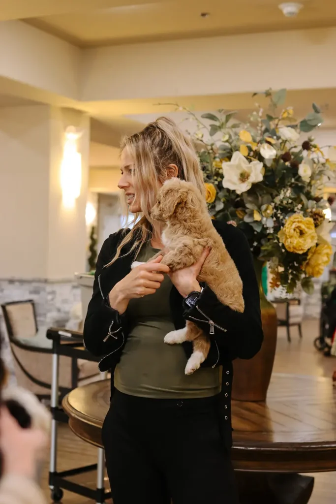 A woman stands indoors holding a small, light brown Doodle puppy near a table with a large floral arrangement in the background, showcasing the charm of Ranch House Doodles.