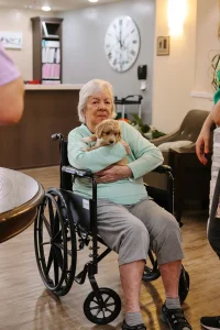 An elderly woman sits in a wheelchair indoors, holding a small Doodle puppy in her lap, with people standing nearby from Ranch House Doodles.