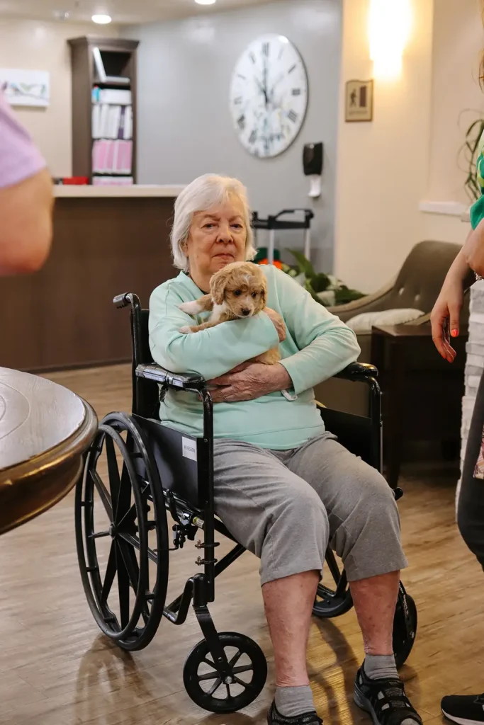 An elderly woman sits in a wheelchair indoors, holding a small Doodle puppy in her lap, with people standing nearby from Ranch House Doodles.
