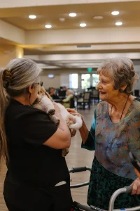 Two elderly women interact while one holds a small dog—possibly one of the Ranch House Doodles puppies—in her arms inside a well-lit communal room with people seated in the background.