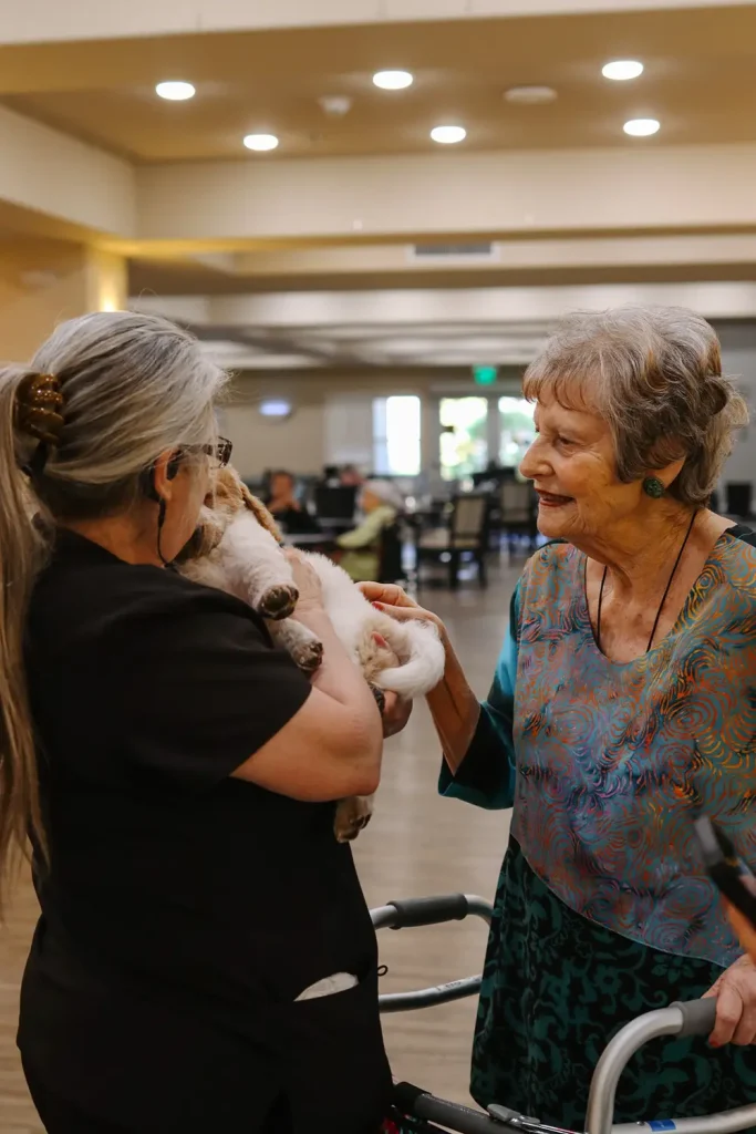 Two elderly women interact while one holds a small dog&mdash;possibly one of the Ranch House Doodles puppies&mdash;in her arms inside a well-lit communal room with people seated in the background.