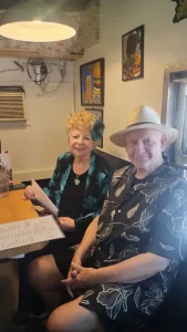 An older woman and man sit at a restaurant table, smiling and enjoying a vibrant community lifestyle. The woman holds a menu and wears a floral accessory; the man sports a patterned shirt and straw hat.