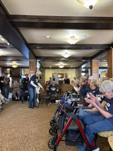 A group of seniors enjoy a vibrant community lifestyle in a cozy room—some using walkers, while one person stands in front as others watch and applaud.