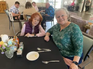 Two elderly women sit at a dining table in a communal area, smiling at the camera, enjoying a warm and welcoming community lifestyle. Other people are seated at tables in the background.