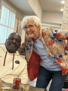 An older man in a white hoodie sits at a table with drinks while an older woman in a colorful jacket leans in beside him, both smiling indoors, capturing a warm moment of lifestyle and community.