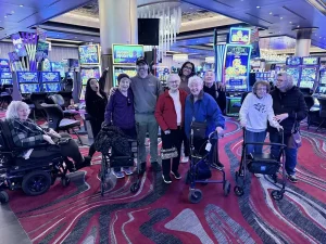 A group of people, embracing community and diverse lifestyles, pose together in a casino with slot machines in the background—some using walkers and wheelchairs.