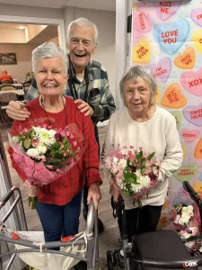 Three older adults, two women holding bouquets and one man standing behind them, smile in front of a Valentine-themed backdrop with conversation heart decorations, celebrating community and lifestyle together.