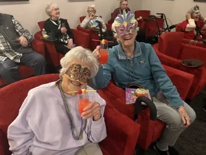 Two elderly women in masks hold colorful drinks and smile in a room with other seniors seated in red chairs, reflecting a vibrant community lifestyle at a festive gathering or party.